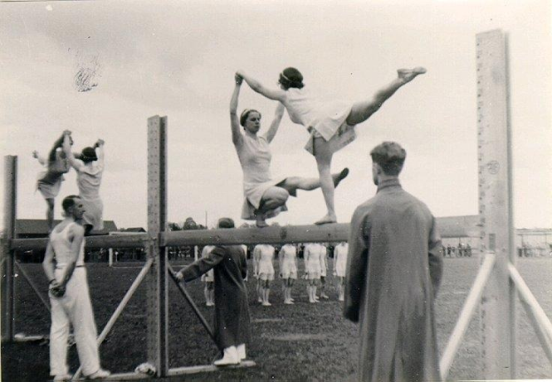 Schwarzes und weißes Foto von Turnerinnen auf einem Balancebalken mit Zuschauern und einer Stadtlandschaft im Hintergrund.