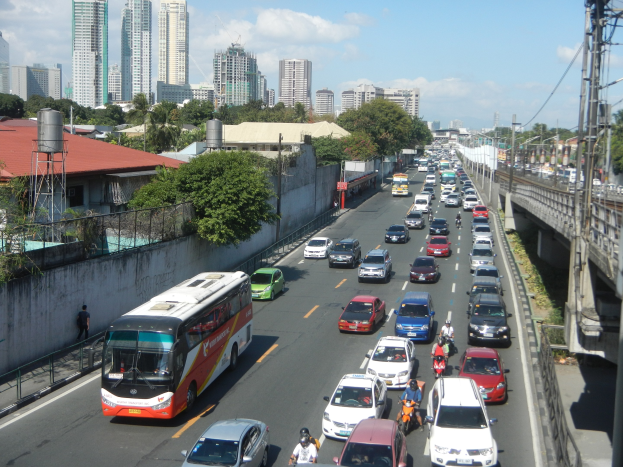 Stadtstraße mit starkem Verkehr von Autos, Bussen und Motorrädern, mit einer Brücke mit Strommasten und -leitungen, Gebäuden, Bäumen und einem bewölkten Himmel im Hintergrund.