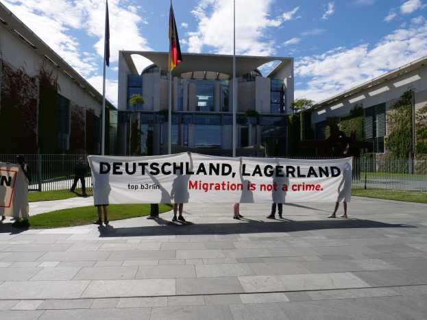 Eine Gruppe von Menschen mit einem Banner, auf dem "Deutschland, Lagerland Migration ist kein Verbrechen" steht, vor einem Gebäude mit Fenstern, umgeben von Gras, einem Metallzaun, Pflanzen, Bäumen und Fahnenstangen unter einem bewölkten Himmel.