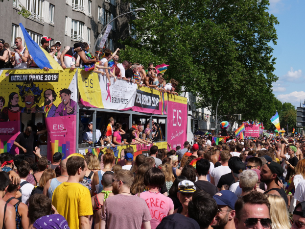 Eine große Menschenmenge marschiert während des Berlin Pride 2019 die Straße entlang, viele tragen Kappen und Schutzbrillen, einige halten Fahnen, mit Bannern, Laternenmasten, Bäumen und Gebäuden im Hintergrund unter einem bewölkten Himmel.