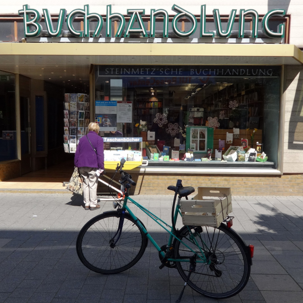 Eine Frau steht neben einem Fahrrad, das vor einem Laden geparkt ist, mit einem Fahrradrad im Vordergrund und durch die Gläser des Ladens sichtbaren Büchern.