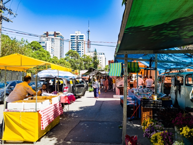 Ein belebter Straßenmarkt mit Menschen, die an parkenden Autos vorbeigehen, Zelte auf beiden Seiten mit Speisen und anderen Artikeln zum Verkauf und einem Hintergrund mit Bäumen, Gebäuden, Strommasten, Drähten, einem Turm und Himmel.
