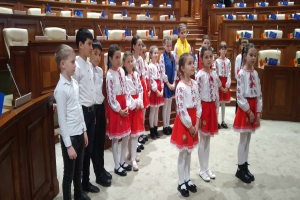 Eine Gruppe von Kindern in weißen und roten Kleidern steht zusammen in einem Raum, mit einem Podium und Mikrofon auf der linken Seite und Stühlen und Tischen mit verschiedenen Gegenständen im Hintergrund, tragend Fahnen, die die Teilnahme an einer Nationalversammlung anzeigen.