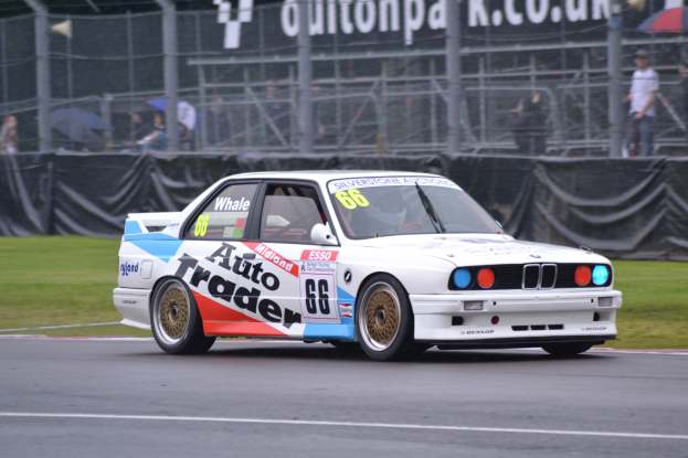 Ein weißer Rennwagen mit Text und Zahlen darauf fährt auf einer Strecke, umgeben von Gras, mit Zuschauern hinter einem Maschendrahtzaun, einige halten Schirme, und eine Fahne im Hintergrund.