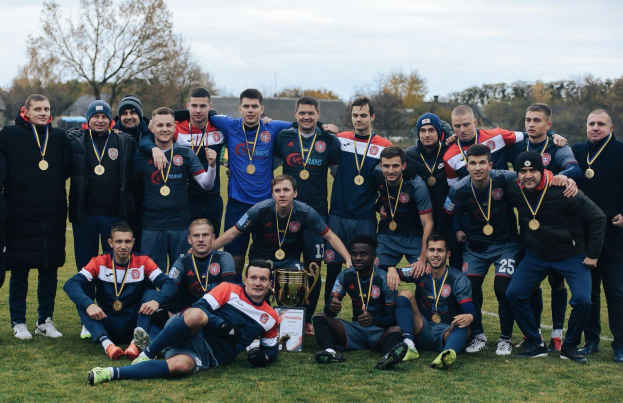 Eine Gruppe von Männern in Sportkleidung steht auf einem Fußballfeld, trägt Medaillen und hält einen Pokal, mit Bäumen und einem klaren blauen Himmel im Hintergrund.