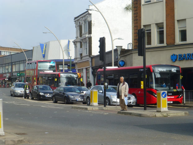 Ein roter Doppeldeckerbus fährt auf einer von hohen Gebäuden gesäumten Stadtstraße, umgeben von Fußgängern, Radfahrern und Ampeln.