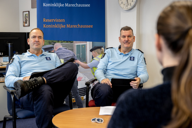 Zwei Polizeibeamte sitzen an einem Tisch mit Papieren, unterhalten sich mit einer Frau, während eine wandmontierte Uhr und ein Banner sowie ein zweiter Tisch mit einem Monitor und Tastatur im Hintergrund zu sehen sind.