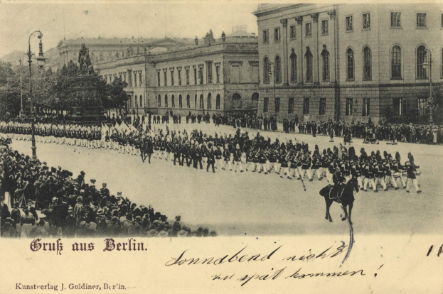 Schwarzes und weißes Foto einer historischen Parade in Berlin, das Menschen zu Fuß und zu Pferd, eine Statue auf einem Sockel, Straßeninfrastruktur, Gebäude und einen bewölkten Himmel zeigt, mit Text unten.