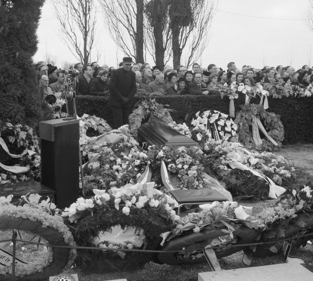Schwarzes und weißes Bild einer Trauerprozession mit einem Mann in einer Mütze, der auf einem Podium spricht, umgeben von Blumen, Stühlen und einer Menge, mit Bäumen und einem klaren Himmel im Hintergrund.