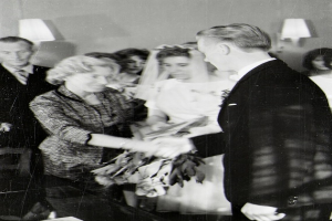 Ein Schwarz-Weiß-Foto eines Brautpaares beim Austausch der Eheringe, wobei die Braut ein weißes Kleid und einen Schleier trägt und einen Strauß hält, und der Bräutigam einen Anzug und eine Krawatte trägt, mit Wandlampen im Hintergrund.