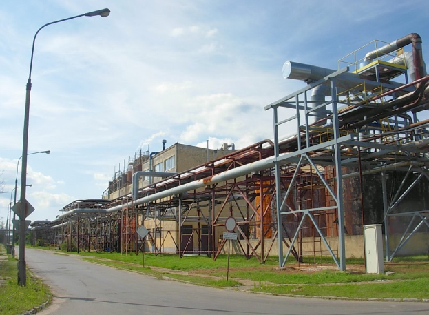 Großes industrielles Fabrikgebäude mit ausgedehnten Rohrleitungen, umgeben von städtischen Elementen einschließlich Gebäuden, Straßeneinrichtungen, Grünflächen und einem bewölkten Himmel.