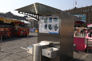 Ein Edelstahl-Wasserbrunnen in der Mitte eines Stadtplatzes, umgeben von Gebäuden, Bäumen, Polen, Fahnen, Werbetafeln, Fahrzeugen, Menschen und anderen Gegenständen, mit dem Himmel oben sichtbar.