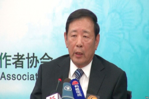 Ein Mann im Anzug sitzt an einem Tisch mit Mikrofonen, während er auf einer Pressekonferenz spricht, mit einem Banner im Hintergrund.