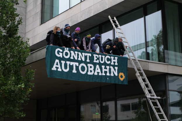 Gruppe von Menschen auf einem Gebäude mit einem Banner, mit einem Baum links und Glasfenstern im Hintergrund, die scheinbar für die deutsche Regierung protestieren.