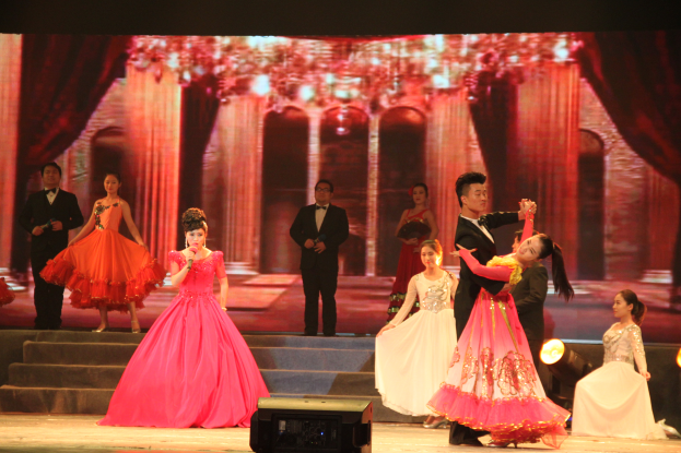 Eine Gruppe von Menschen auf einer Bühne, beleuchtet von Scheinwerfern, mit Stufen und einem Bildschirm im Hintergrund und einem Lautsprecher unten, was eine Tanzvorstellung suggeriert.