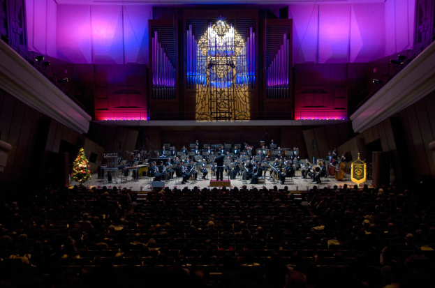 Großer Saal mit Publikum bei einem Konzert auf der Bühne, mit einem Weihnachtsbaum und dekorierten Wänden mit Lichtern.