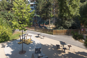 Ein Park mit Bänken, Tischen, Bäumen, Pflanzen, Gras, einer Straße, einem Zaun, ein paar Menschen, Gebäuden im Hintergrund und einem klaren blauen Himmel.