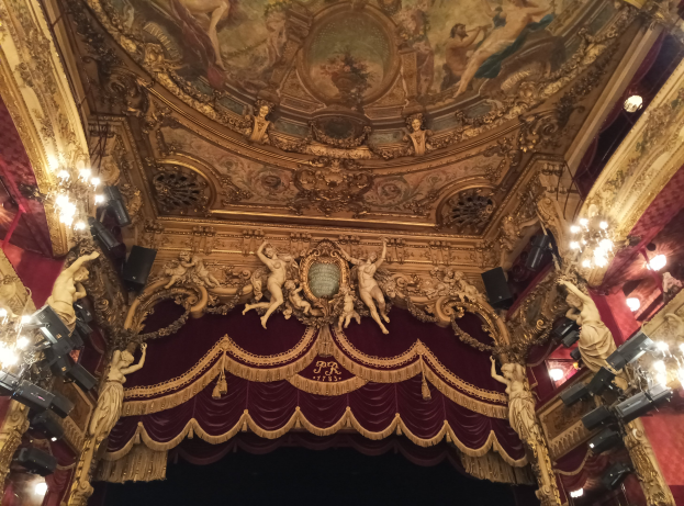 Theaterinnenraum mit roter Gardine, Kronleuchtern, Skulpturen, Deckenleuchten und einem Gemälde, das eine Bühne mit einigen Menschen am Boden zeigt.