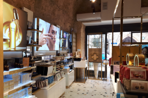 Innenansicht eines Kosmetikladens mit Regalen voller verschiedener Schönheitsprodukte, wandmontierten Bildschirmen, Deckenlampen, einer Klimaanlage und Glastüren, die eine Person, ein Auto und ein Gebäude zeigen.