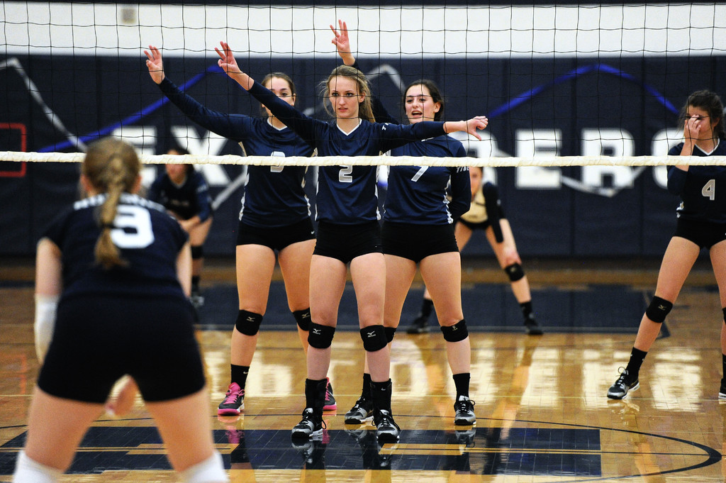 Drei Mädels bei einem Volleyballspiel auf dem Feld mit erhobenen Händen, ein Netz davor und ein Zaun im Hintergrund.