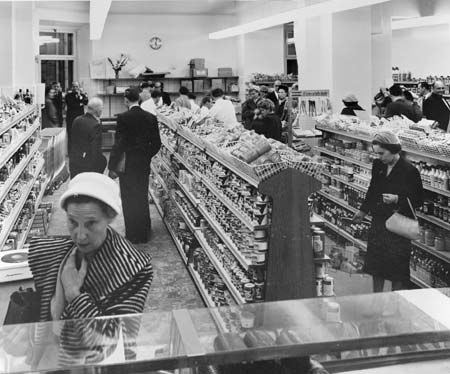Schwarzes und weißes Foto von Menschen beim Einkaufen in einem Supermarkt mit Lebensmittelregalen, einige halten Taschen, eine Uhr an der Wand und Deckenlampen.