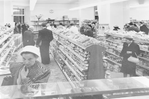 Schwarzes und weißes Foto von Menschen beim Einkaufen in einem Supermarkt mit Lebensmittelregalen, einige halten Taschen, eine Uhr an der Wand und Deckenlampen.