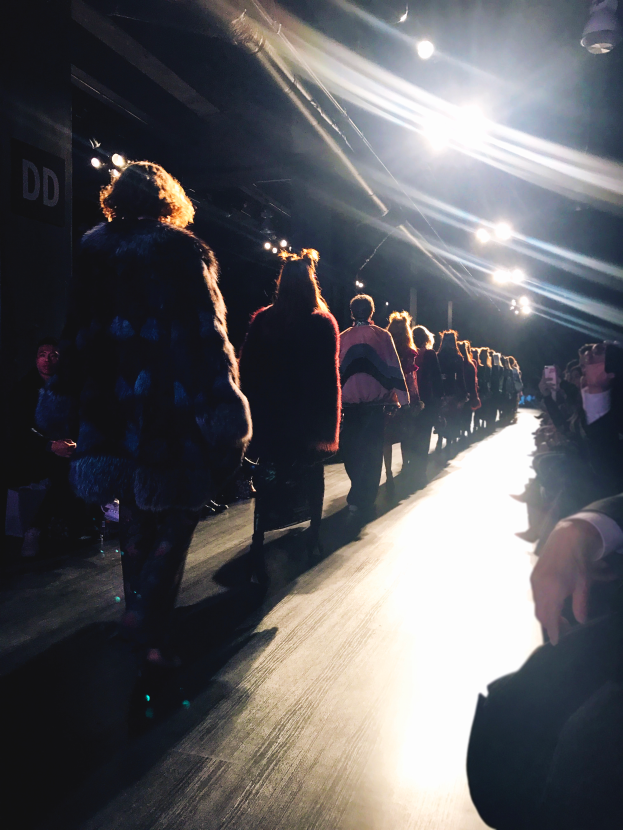 Eine Gruppe von Menschen läuft auf einem Laufsteg bei einer Fashion Show, beleuchtet von hellen Scheinwerfern, während Zuschauer auf beiden Seiten das Spektakel beobachten.