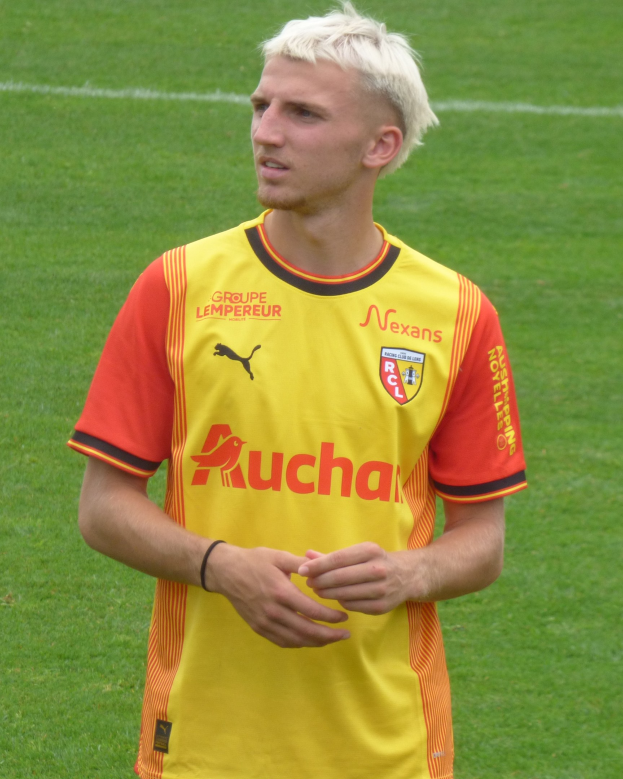 Ein Mann in einem gelben und orangefarbenen Fußballtrikot steht selbstbewusst auf einem Fußballfeld und sieht aus wie ein Flügelspieler mit entschlossener Miene.