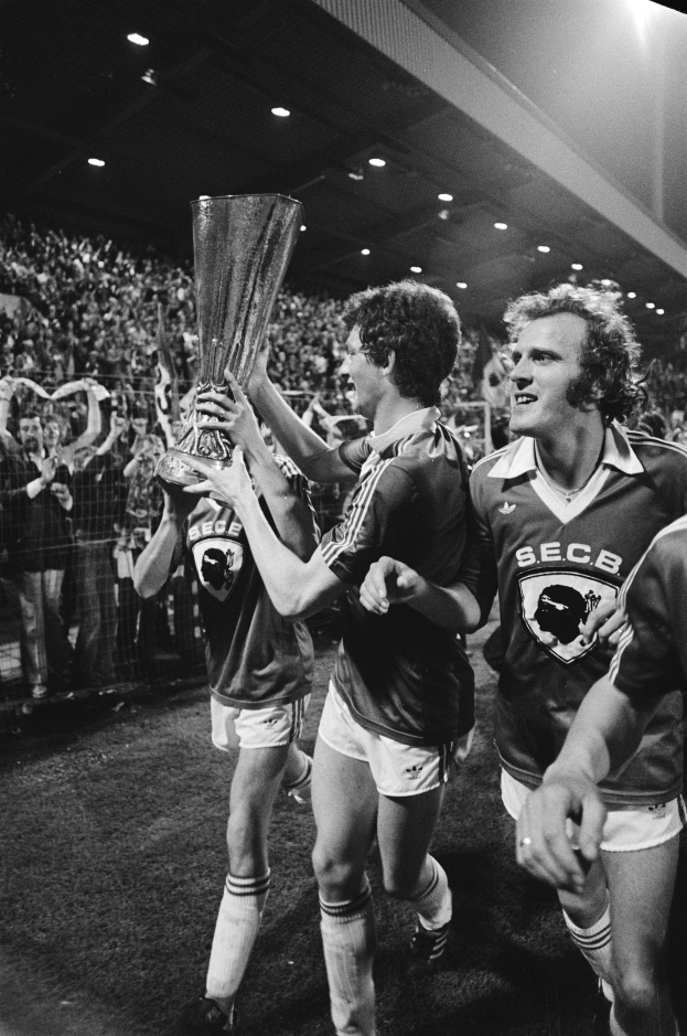 Schwarzes und weißes Bild von Fußballspielern, die mit einem Pokal auf einem Feld feiern, umgeben von einer Menge hinter einem Zaun unter Stadionbeleuchtung.