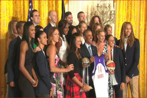 Präsident Obama und First Lady Michelle Obama posieren mit dem Frauen-Basketball-Team im Oval Office des Weißen Hauses, mit einem Basketball und einer Trophäe, einem Flagge, Vorhängen und einem Kerzenleuchter im Hintergrund.