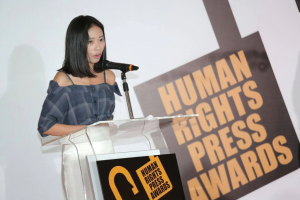 Eine Frau in einem schwarzen Kleid steht an einem Podium mit einem Mikrofon und hält eine Rede oder Präsentation, mit einem Banner dahinter, das "Human Rights Press Awards" lautet.