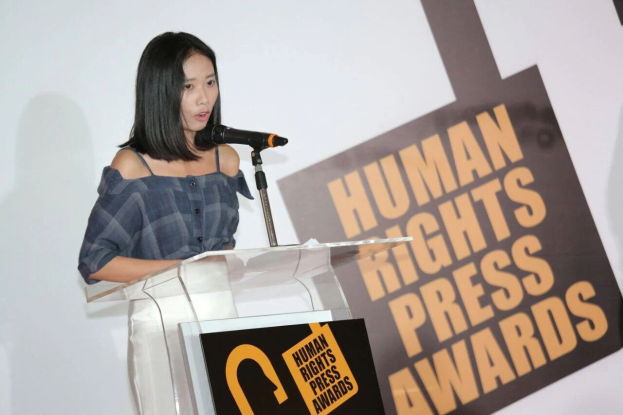 Eine Frau in einem schwarzen Kleid steht an einem Podium mit einem Mikrofon und hält eine Rede oder Präsentation, mit einem Banner dahinter, das "Human Rights Press Awards" lautet.