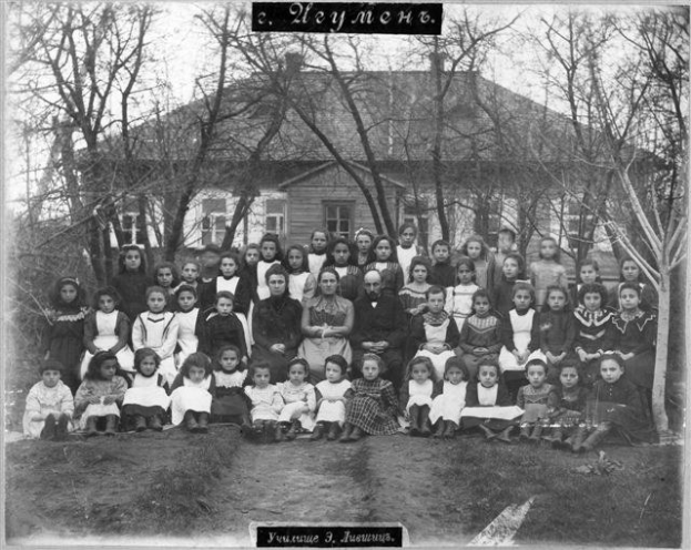 Schwarzes und weißes Foto einer Gruppe von Kindern, die vor einem Fensterbau mit Text posieren, umgeben von Bäumen und einem klaren Himmel.