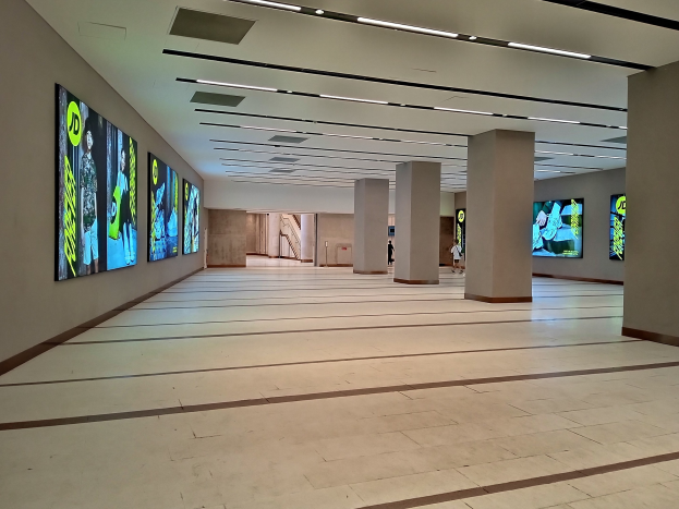 Ein großer Raum mit zahlreichen wandmontierten Fernsehern, beleuchtet von Deckenlampen, mit ein paar Menschen und sichtbarem Boden.