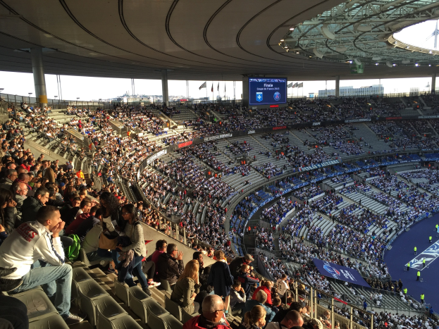 Eine große Menschenmenge sitzt im Allianz Stadion in München, Deutschland, und schaut ein Fußballspiel. Auf der rechten Seite befindet sich eine Bühne und im Hintergrund sind Fahnen, Stangen und ein Bildschirm zu sehen.