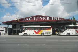 Zwei Busse vor einem Gebäude mit einer 'Jac Liner Inc.'-Plakette geparkt, mit Menschen auf dem Gehweg, Strommasten mit Drähten, Bäumen und einem bewölkten Himmel im Hintergrund.