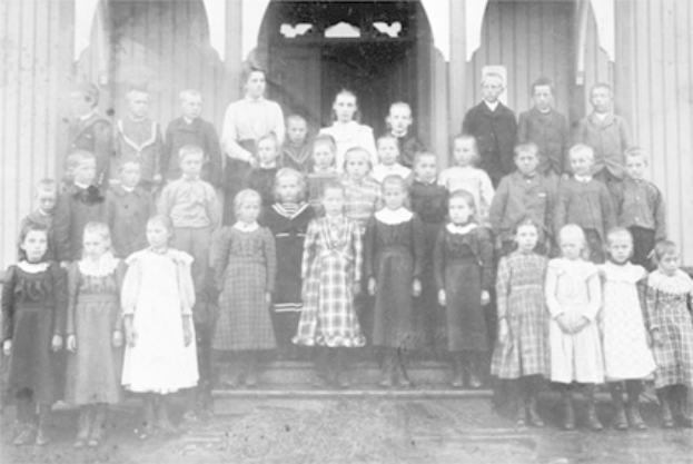 Ein Schwarz-Weiß-Foto einer Gruppe von Kindern, die vor einem Gebäude mit einer Tür und Säulen stehen, mit der Beschriftung "Erste Grundschule" unten.