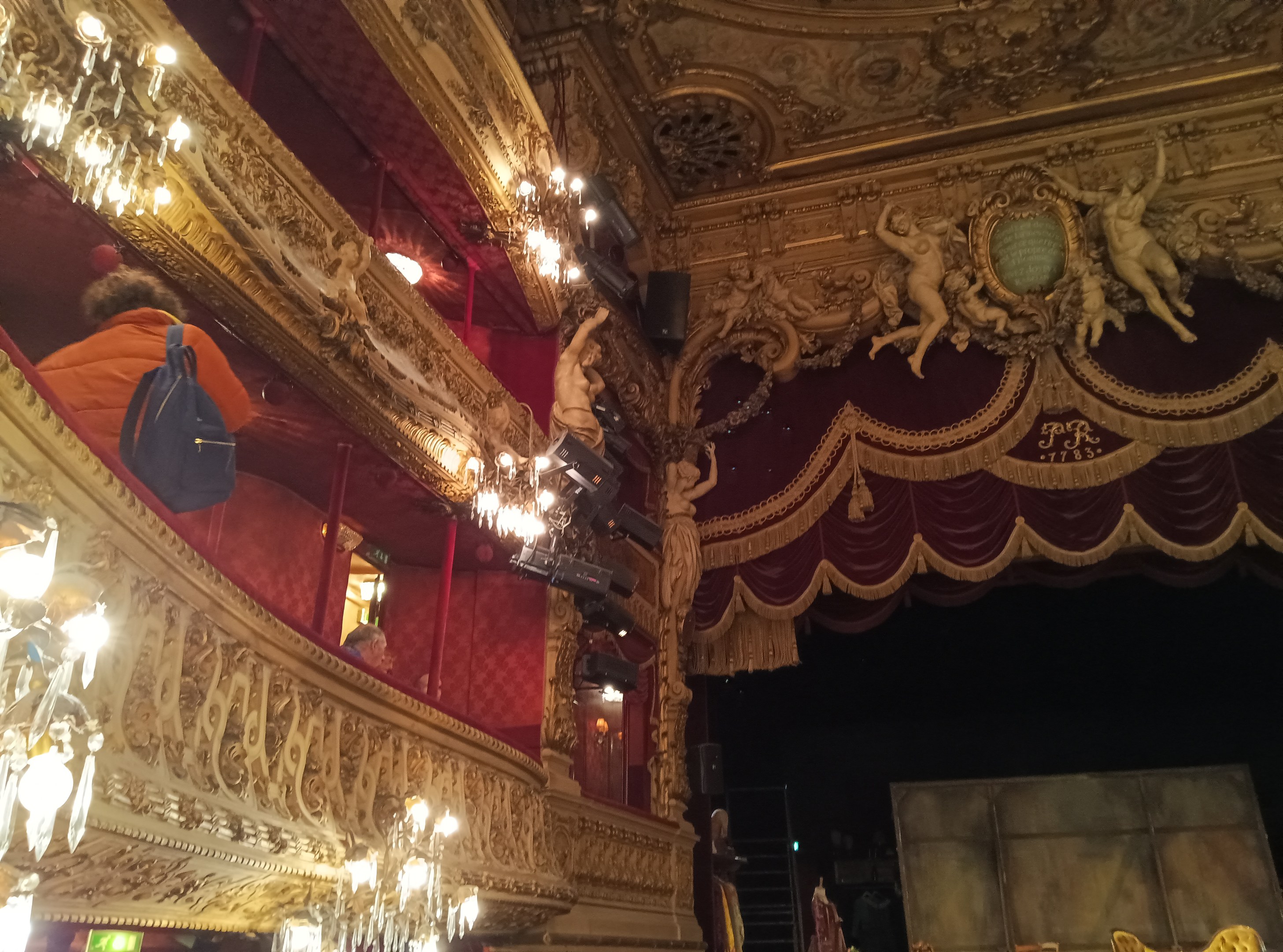 Das Innere eines Theaters mit Kronleuchtern an der Decke, Skulpturen an den Wänden und Menschen auf der Bühne, mit verstreuten Gegenständen, die eine lebhafte Atmosphäre schaffen.