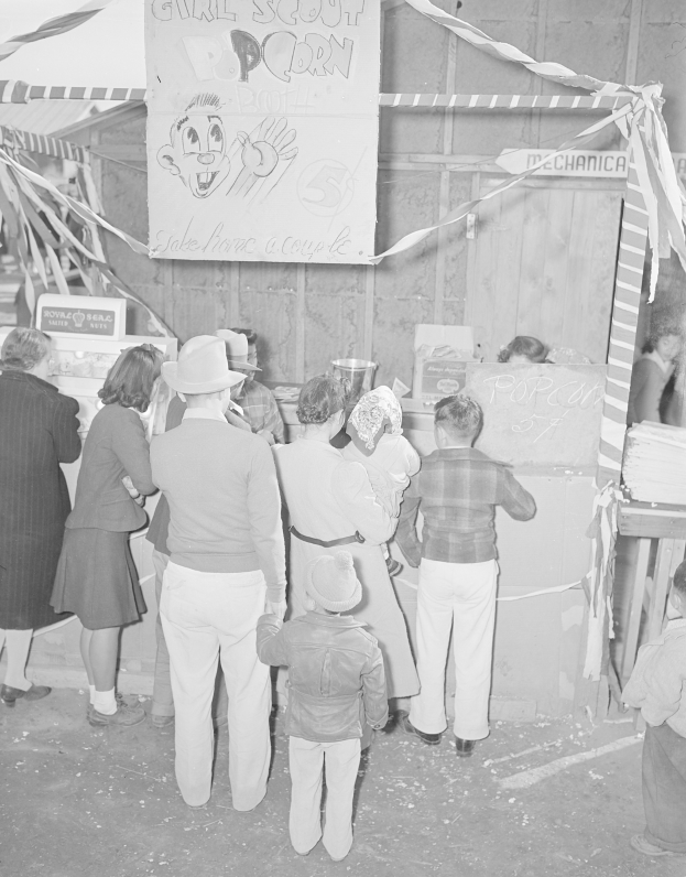 Schwarzes und weißes Bild einer Gruppe von Menschen, die vor einem Karnevalsstand mit Bändern und einer Schautafel stehen.