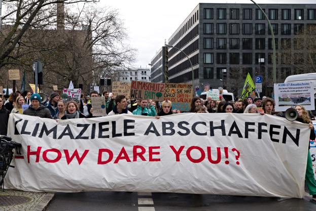Gruppe von Menschen geht eine Straße entlang und hält ein Transparent mit der Aufschrift "Klimaziele Abschaffen - Wie wagst du es?" mit Fahrrädern auf der linken Seite, Bäumen, Gebäuden, Laternenmasten und einem klaren blauen Himmel im Hintergrund.