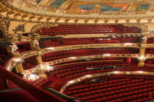 Ein Blick hinter die Kulissen der Opernhaus-Bühne im Londoner Royal Opera House, mit Sitzreihen, Geländern, Säulen, Deckenleuchten, Wandskulpturen und einer Deckenmalerei.