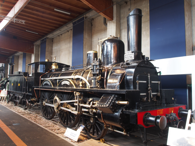 Eine gro├če, alte Dampflokomotive, die in einem Museum ausgestellt ist, auf Eisenbahnschienen, mit informativen Schildern, St├Ąnden und anderen Ausstellungsst├Ącken in der N├Ąhe, vor einer Wand mit Deckenleuchten.