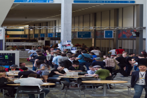 Ein belebter Flughafenvorplatz mit wartenden, arbeitenden und verschiedenen Aktivitäten nachgehenden Menschen, Richtungsschilder an der rechten Wand und darüber sowie an einer Eisenstange an der Decke befestigte Lampen.