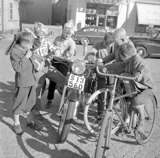 Eine Gruppe von Kindern umringt ein Motorrad auf einer Straße mit Gebäuden, einem Zaun, einem Schild und einem bewölkten Himmel in Schwarzweiß; ein Kind sitzt auf dem Motorrad.