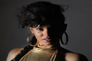 Eine Frau in einem goldenen Kleid und großen Ohrringen steht selbstbewusst vor einer Wand, ihr glatter Bob-Haarschnitt und ihr natürliches Make-up unterstreichen ihren entschlossenen Gesichtsausdruck, während sie direkt in die Kamera blickt.