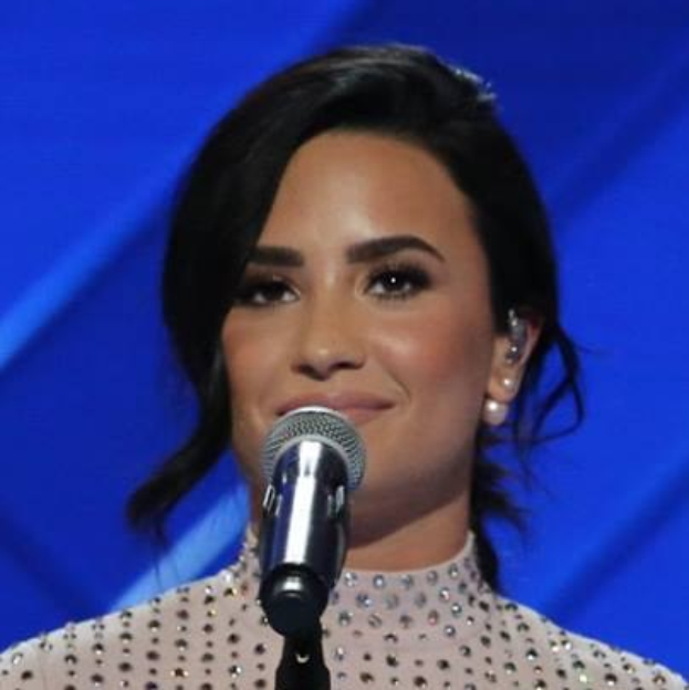 Demi Lovato steht mit einem Mikrofon auf der Bühne der Grammys, trägt ein schwarzes Kleid und einen eleganten Bob und hat einen selbstbewussten Gesichtsausdruck vor einem blauen Hintergrund.