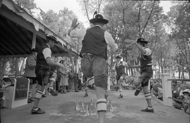 Eine Gruppe von Männern in traditioneller bayrischer Tracht tanzt auf einer Bühne vor einem gemischten Publikum, mit Bäumen und einer Hütte im Hintergrund, dargestellt in Schwarz-Weiß.