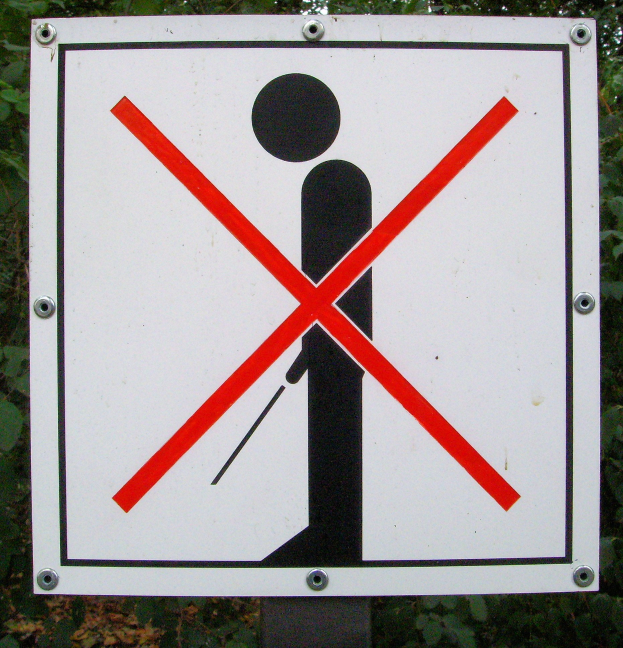 Weißes Parkverbotsschild mit schwarzem Text und rotem Kreuzsymbol auf einem Pfahl montiert, umgeben von Bäumen in einem bewaldeten Gebiet.