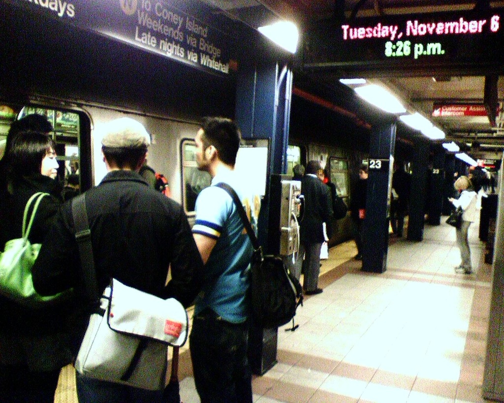 Ein überfüllter Bahnsteig mit Menschen mit Taschen, einem Zug auf der linken Seite und einer Texttafel oben.