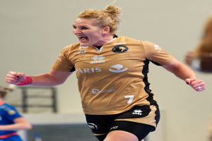 Eine Frau in braunem T-Shirt und schwarzen Shorts rennt auf einem Futsalplatz, feiernd mit triumphaler Miene, während zwei andere Frauen im Hintergrund unscharf sind.